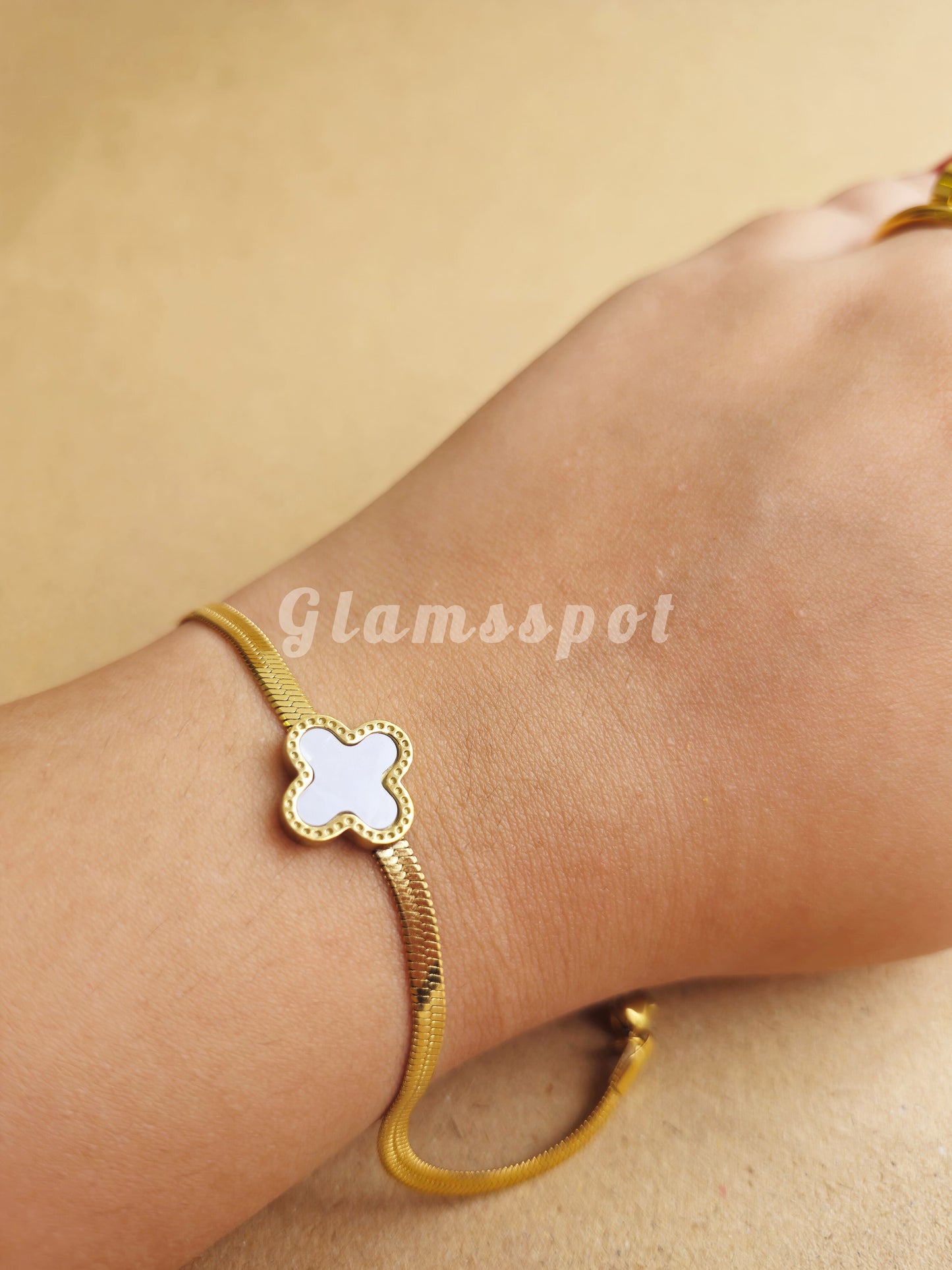 Gold bracelet with a white flower charm on a beige background, branded 'Glamsspot'.