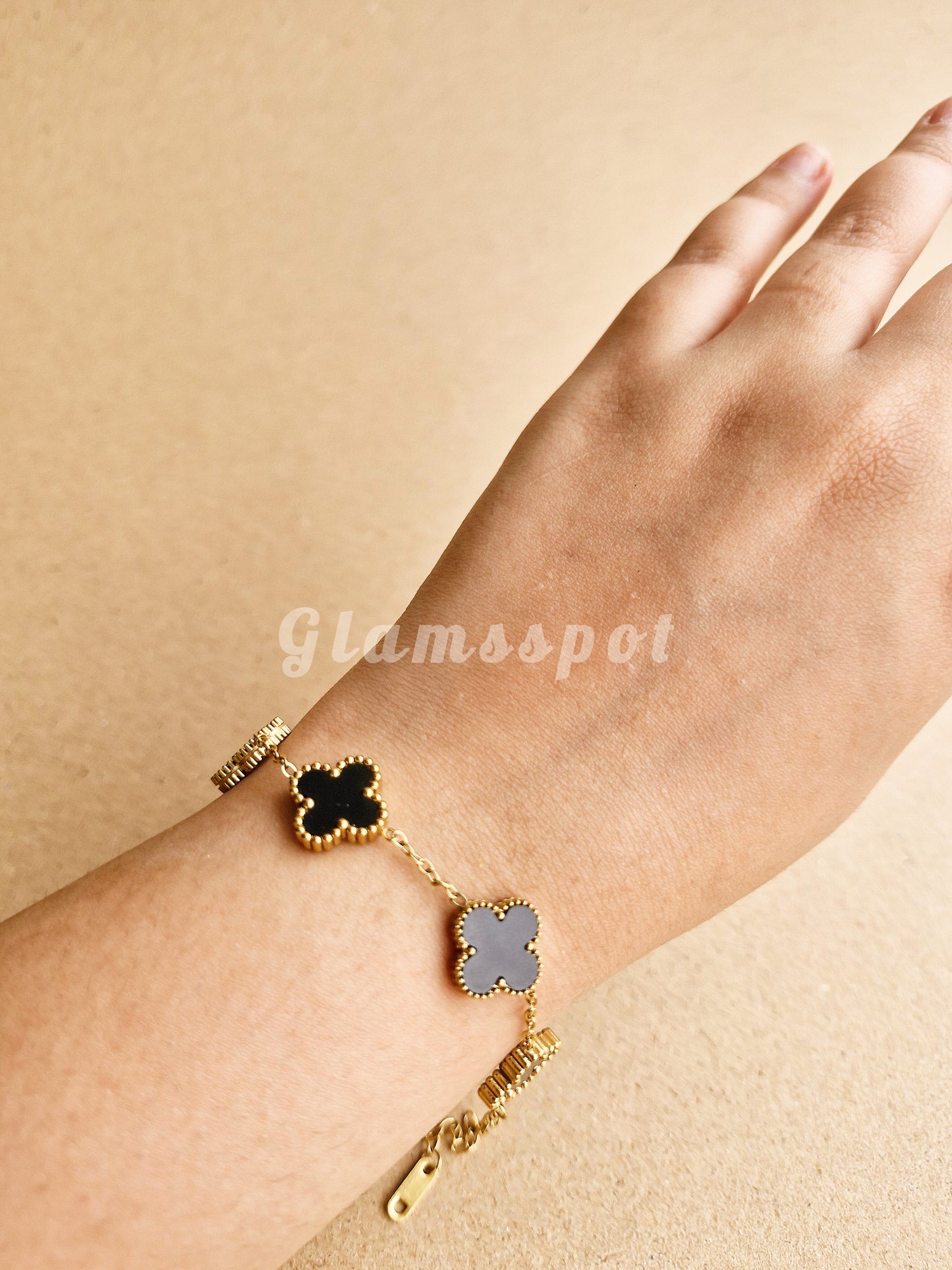 Black Clover Bracelet with clover-shaped charms on a wrist against a beige background, branded 'Glamspot'.