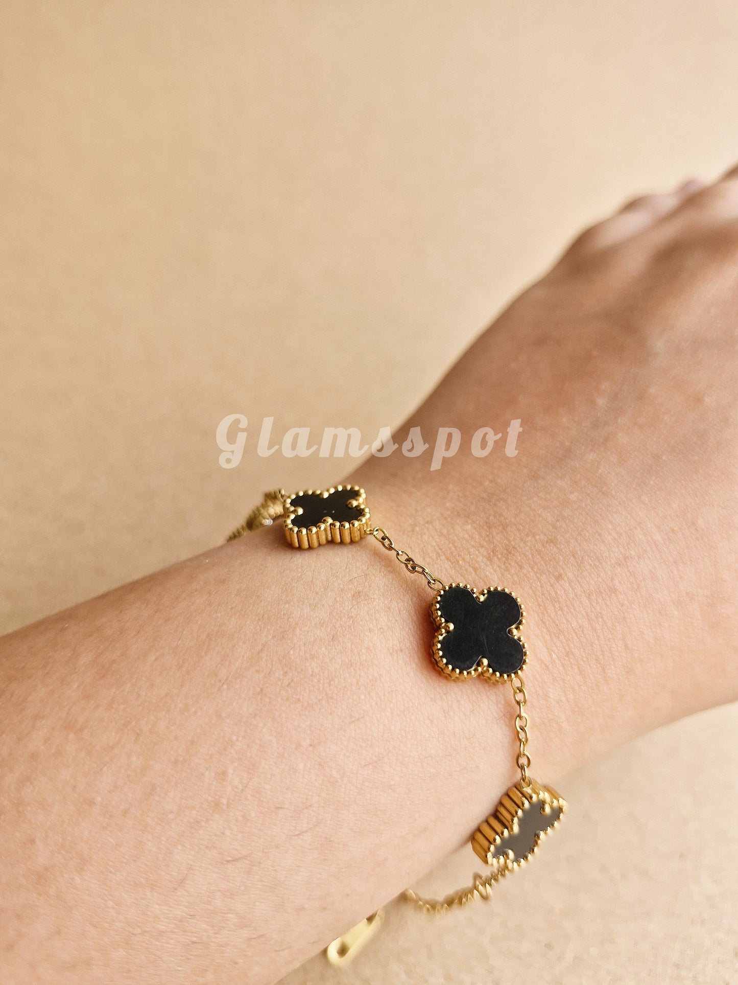 Gold bracelet with black clover-shaped charms on a wrist against a beige background, branded 'Glamispot'.